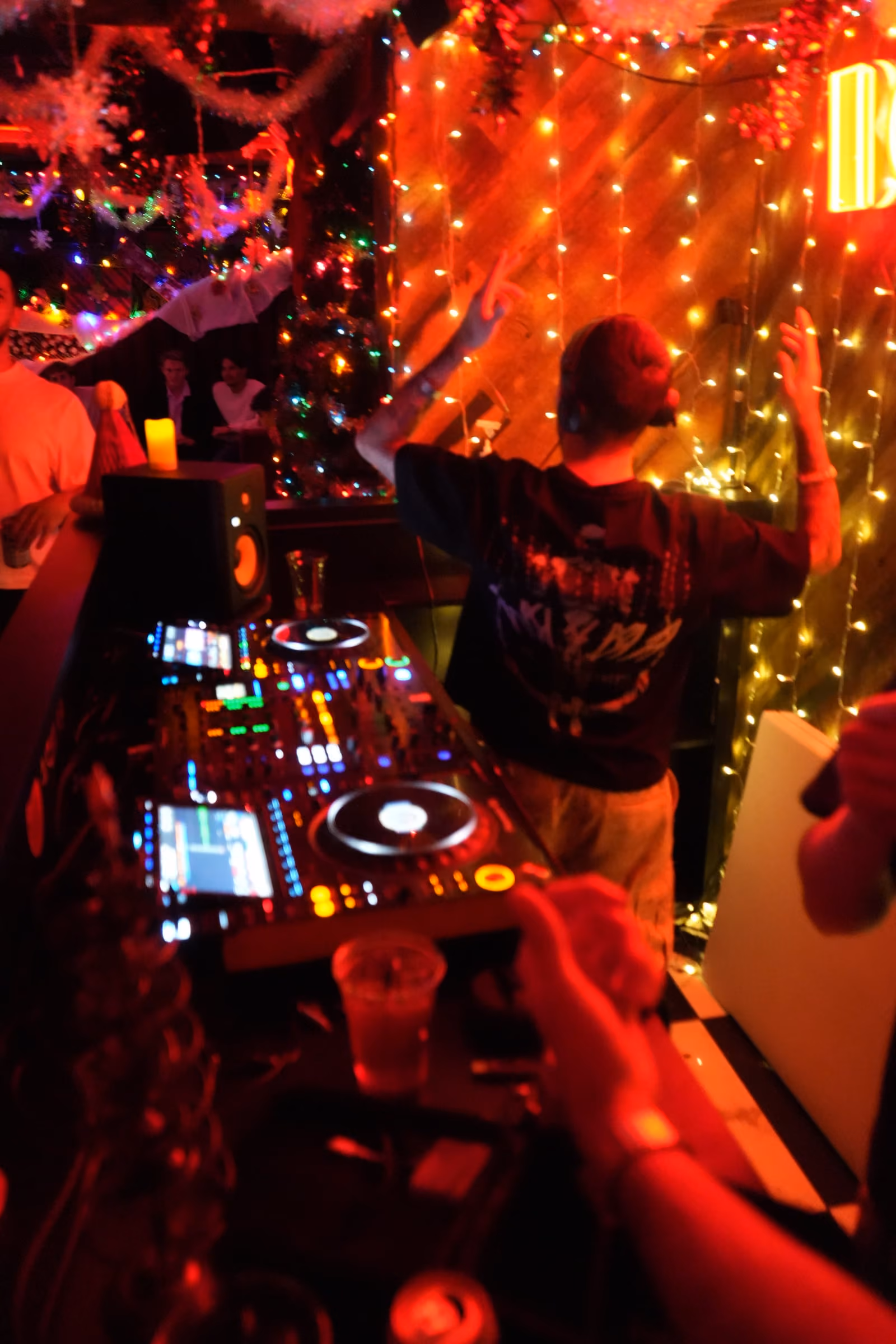AED with arms raised behind the decks in a booth strung with fairy lights